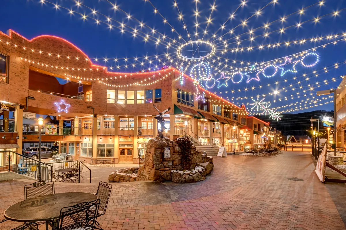 A cheerful exterior view of a festive plaza, with string lights overhead, decorative storefronts, and a central fountain. The scene is alive with people strolling, socializing, dining, and enjoying the magical evening atmosphere.