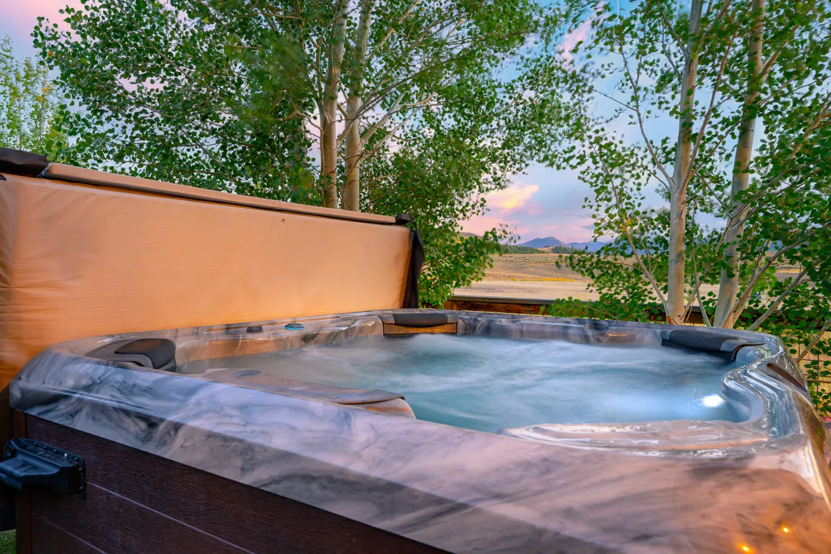 The image captures a hot tub nestled among trees with distant mountain views, emphasizing relaxation and a strong connection to nature. The mood is serene and peaceful, inviting guests to unwind while soaking in the beautiful mountain scenery.