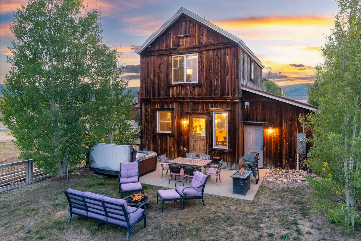 This image displays the exterior of a wooden house featuring a welcoming outdoor seating area, a fire pit, and a jacuzzi hot tub. The setting is perfect for both relaxation and socializing, with the inviting hot tub being a clear highlight. Ambient lighting and sunset sky create a cozy, inviting atmosphere ideal for enjoying the Colorado outdoors.