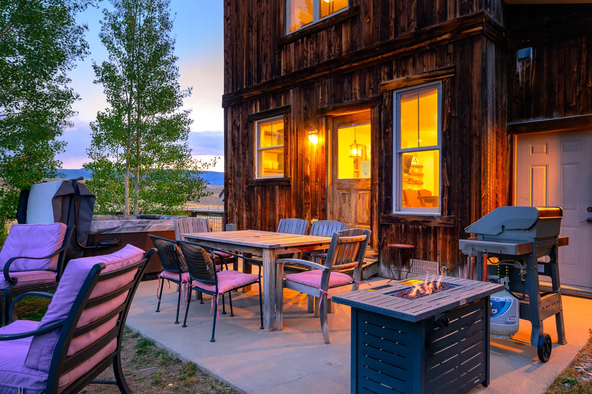 This exterior image features an outdoor dining area with a dining table, fire pit, grill, and comfortable seating. The scene is set for evening gatherings, sunset dinners, and barbecues, with warm lighting creating an inviting ambiance.