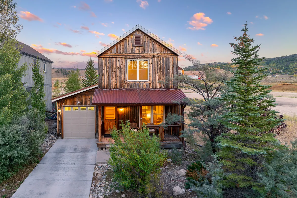 A welcoming exterior view of a wooden house with a front porch, large windows, and a distinctive red metal roof. The porch is ideal for morning coffee or evening relaxation, surrounded by natural beauty.