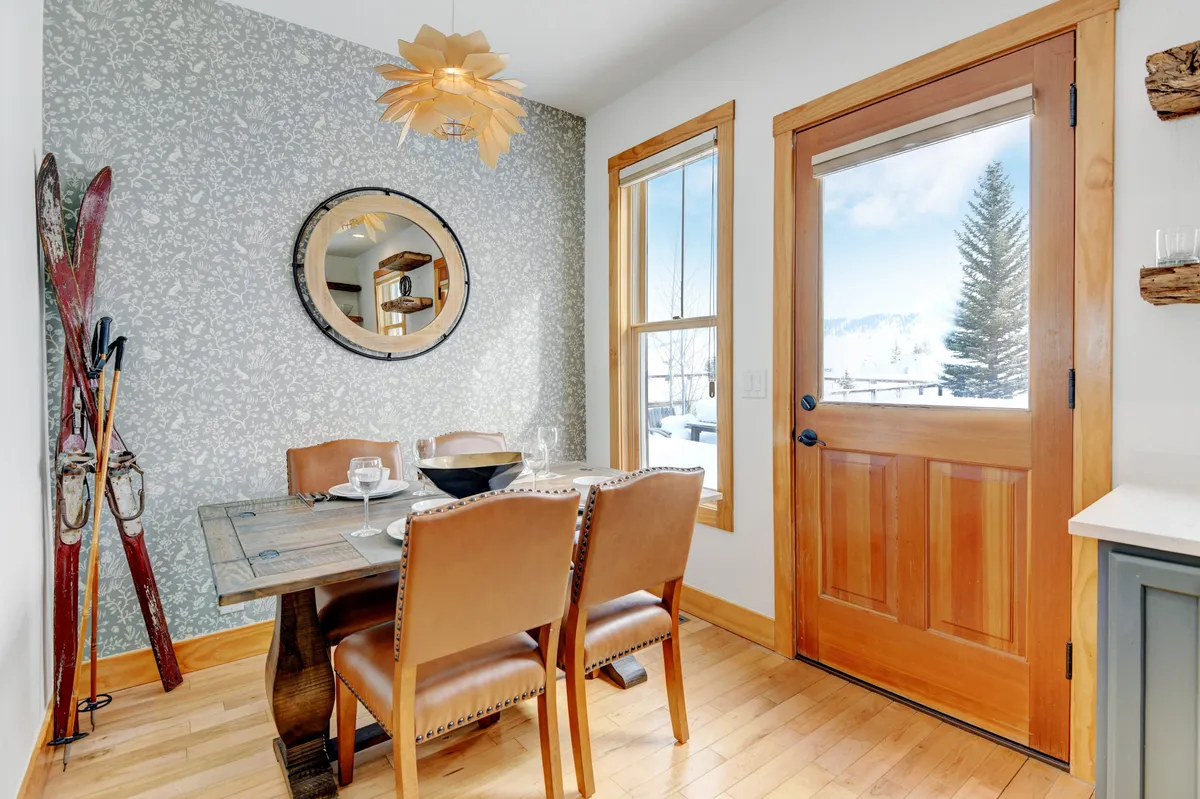 A cozy dining table set near a window with scenic views, complemented by a decorative mirror, unique light fixture, and ski equipment. The setup suggests intimate gatherings, meals, and socializing after outdoor adventures.