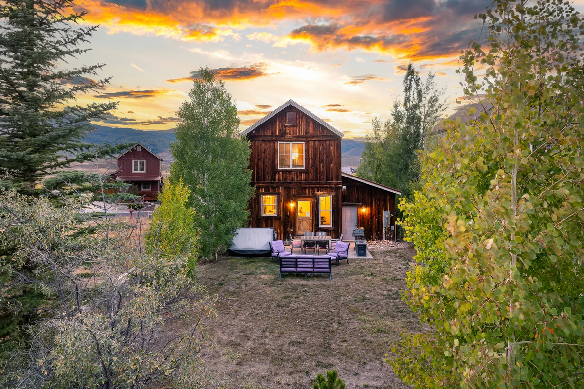 This exterior image showcases the rustic wooden house, a cozy seating area on the porch, and a vibrant sunset in the background. The surrounding landscape hints at nearby trails and open outdoor spaces ideal for walking and exploring with pets.
