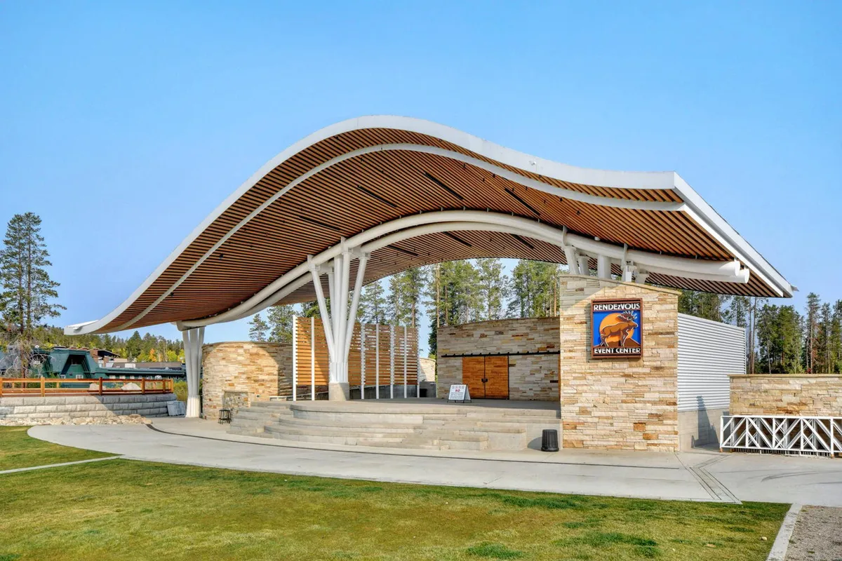 This exterior shot features a modern curved roof structure set amidst stone walls, with a stage area in the foreground. The open-air design hints at a lively gathering space ideal for events and performances. The festive atmosphere is further suggested by visible lighting and arrangement for community use.