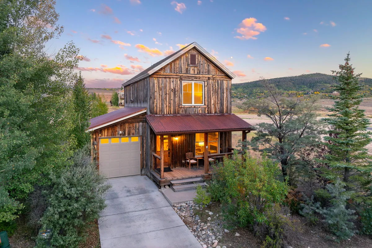 The image showcases the rustic wooden façade of a single-family luxury cabin in Granby, Colorado, with a large inviting front porch and warmly lit windows. The scene evokes a cozy, welcoming mood, suggesting guests relaxing on the porch, enjoying a sunset view, and gathering with loved ones. The setting highlights serene natural surroundings and the peaceful ambiance of the property.