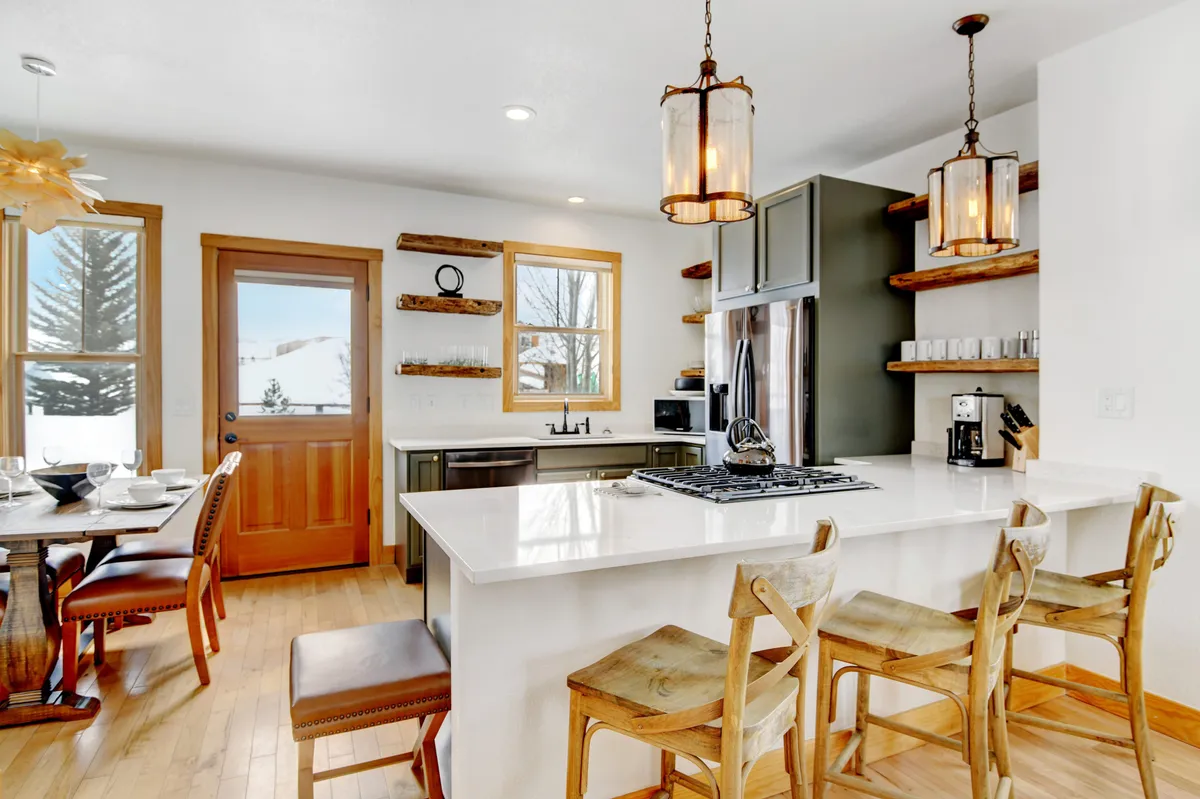 An open-concept kitchen and dining area with a generous kitchen island, dining table set for meals, and large windows that offer natural light and views. Shelves display dishes, and the space is arranged for both cooking and entertaining, showcasing the seamless flow among the kitchen, dining, and living zones.