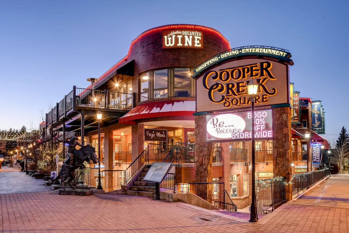 This image features the vibrant entrance to Cooper Creek Square, showcasing its wine store, inviting outdoor seating, decorative lighting, and a bustling, lively atmosphere perfect for shopping, dining, and socializing.