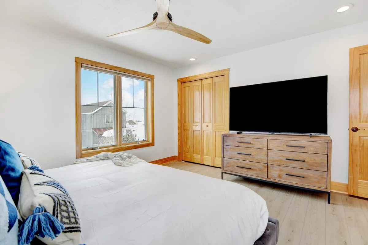 A cozy bedroom with a large window providing a view of the outdoors, a comfortable bed with textured throw pillows, wooden furniture, and a ceiling fan. The space is illuminated with soft, natural light, creating a peaceful retreat atmosphere.