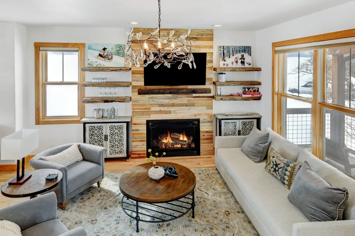 A welcoming living area featuring a prominent fireplace, elegant chandelier, coffee table, and tastefully decorated shelves. The arrangement invites socializing and relaxation, with ample seating and warm lighting that enhances the cozy ambiance. The wall-mounted TV is visible, supporting entertainment and family gatherings.