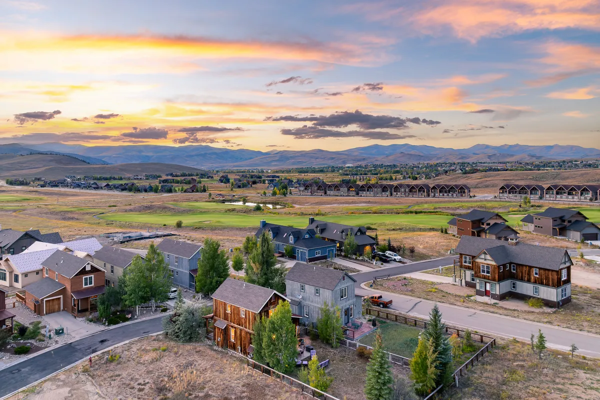 A tranquil, wide-angle view of rolling open fields, a glowing sunset sky, distant mountain ranges, and a few residential homes. The scene invites viewers to imagine hiking nearby trails, golfing, or gathering with family in the great outdoors.